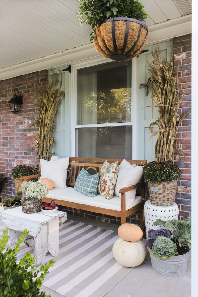 Fall front porch with wooden bench, pillows, cornstalks, and mums