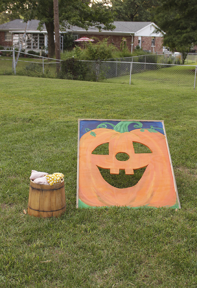 Cornhole board in pumpkin design resting in grass with wooden bucket of bean bags next to it
