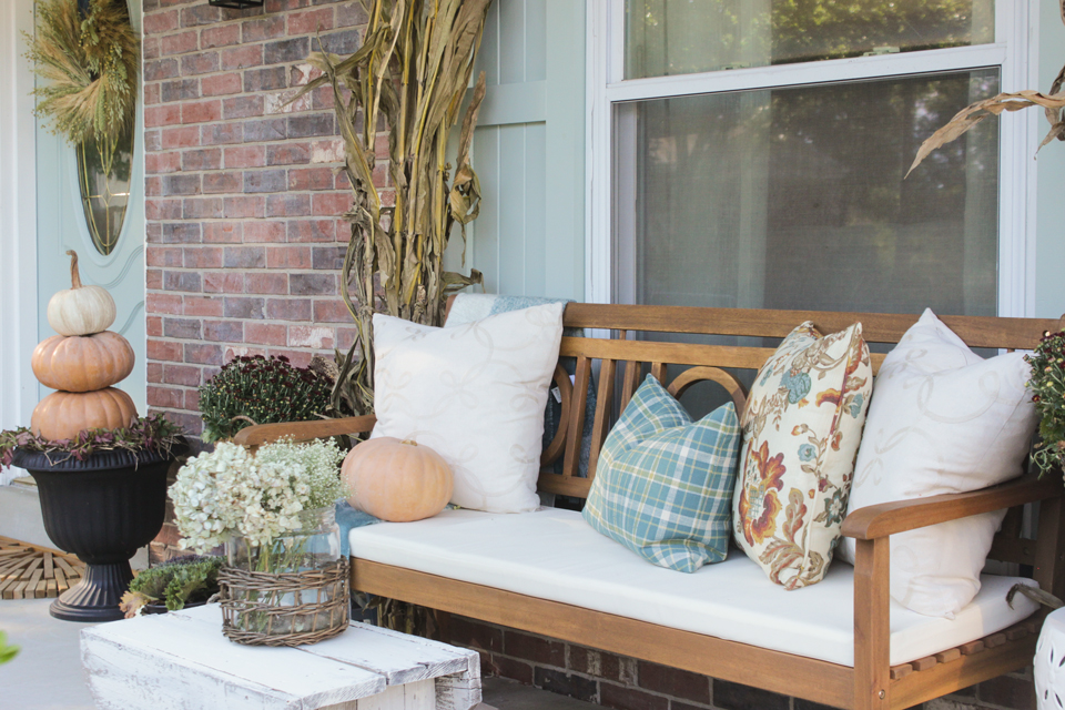 Fall front porch with wooden bench, pillows, cornstalks, and pumpkin topiary